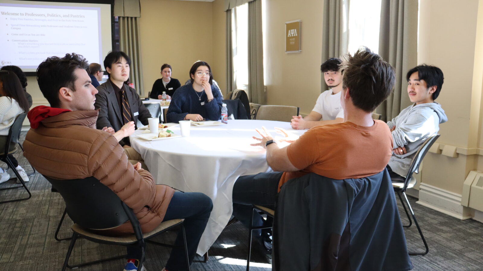 A group of students sits around a round table in a campus meeting room, engaged in conversation at an event called 'Professors, Politics, and Pastries,' as shown on the projection screen in the background. Food and drinks are on the table, and a faculty member with a name badge is visible in the background.