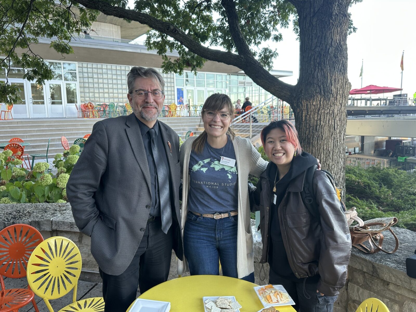 Three people smile for a photo at an outdoor event at the UW–Madison Memorial Union Terrace. The person in the middle wears an 'International Studies Major' t-shirt and a name badge. Colorful patio chairs, a yellow table with snacks, and the Union building are visible in the background.